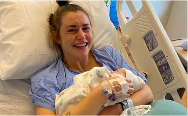 Woman in hospital bed smiling while holding newborn baby wrapped in a blanket.