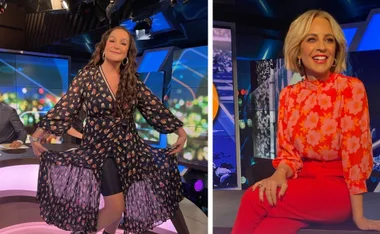 Two women on a TV set, one in a floral dress posing playfully, the other in a bright floral top, smiling brightly.