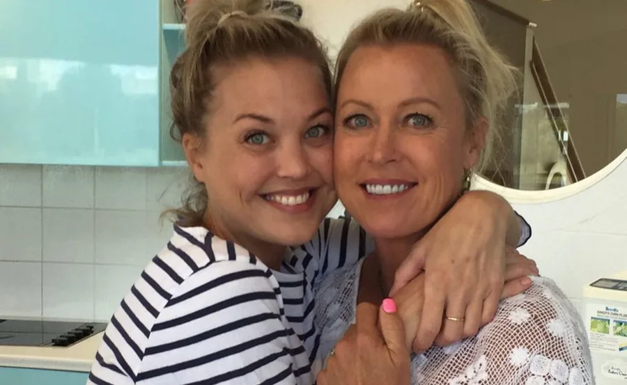 Two women smiling warmly, embracing each other, both with blonde hair and casual attire.