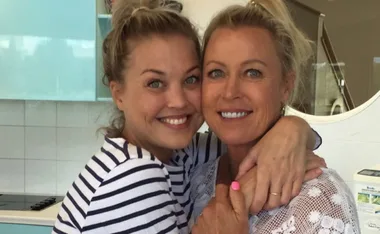 Two women smiling warmly, embracing each other, both with blonde hair and casual attire.
