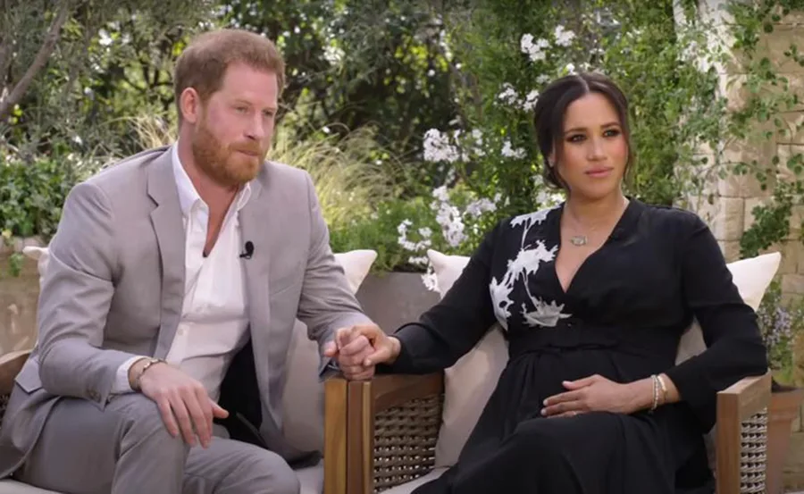 Couple sitting outdoors on chairs, holding hands, amid a garden setting with greenery in the background.