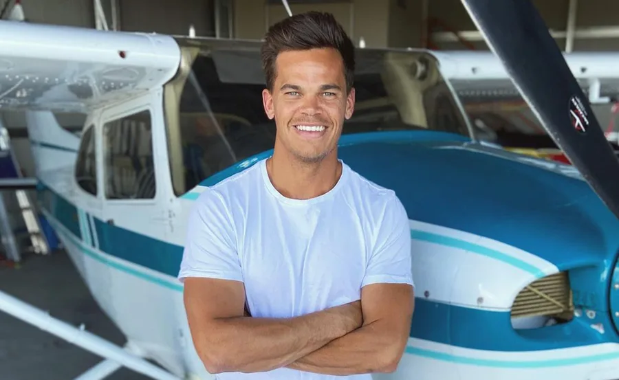 A person in a white shirt stands in front of a small airplane, smiling with arms crossed.