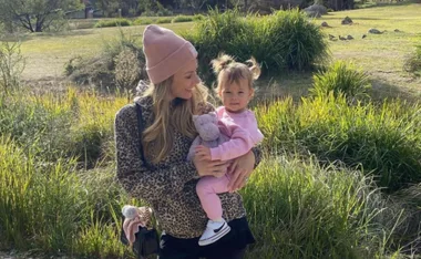 Mother in a beanie holding a child with a toy, outdoors in a grassy area.