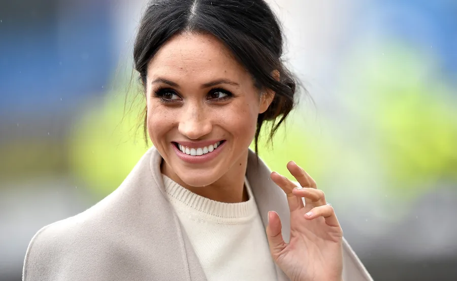 Smiling woman with dark hair in an updo, wearing a light-colored coat, looks to the side and waves.