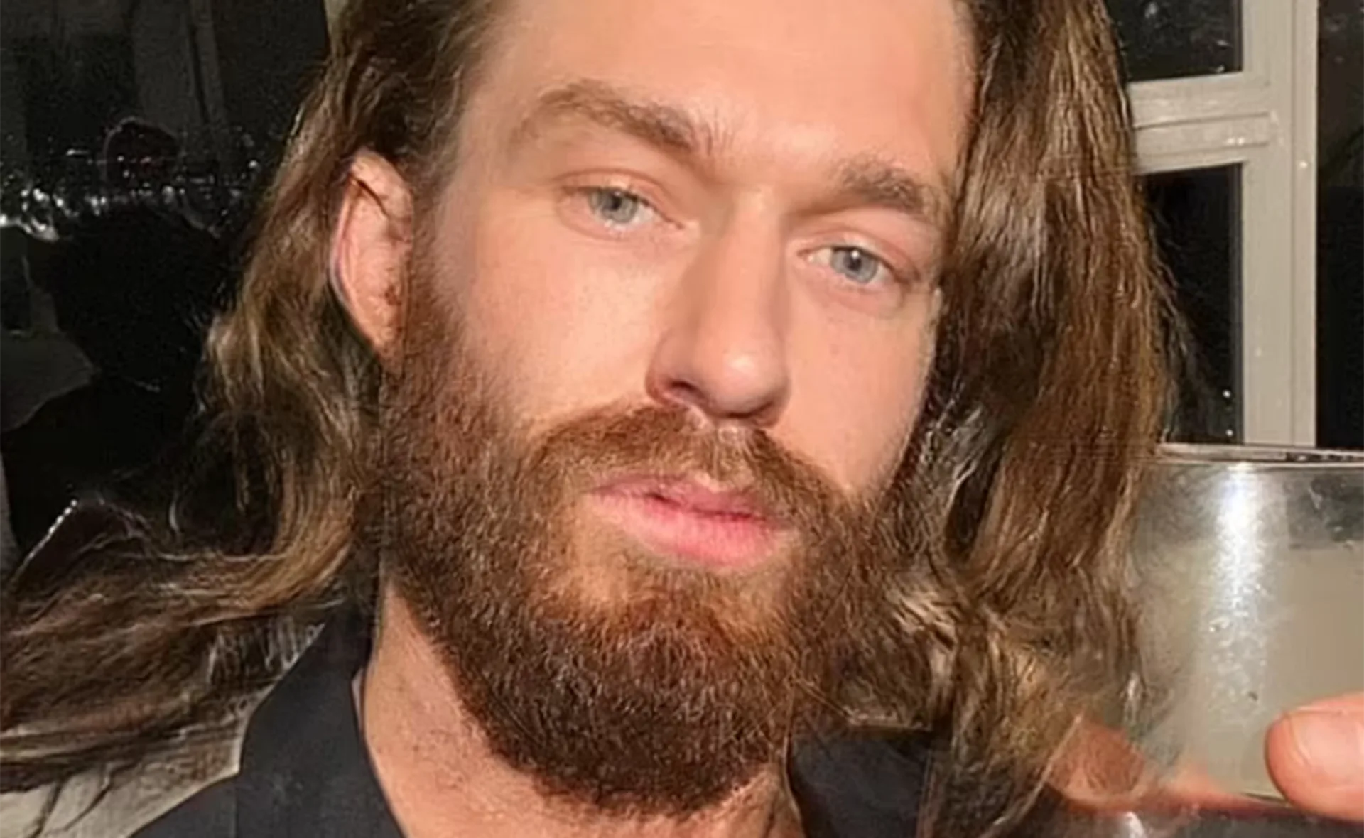 A person with long hair and a beard holding a glass, in a dimly lit room.