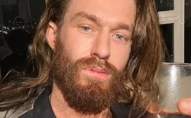A person with long hair and a beard holding a glass, in a dimly lit room.