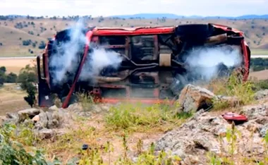 Overturned red car with smoke rising, on rocky terrain with distant hills.