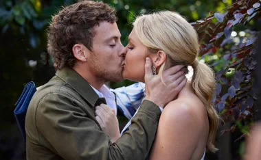 A man and woman share a passionate kiss outdoors, surrounded by lush greenery.