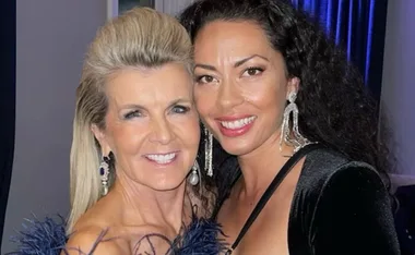 Two women smiling and posing together, wearing elegant outfits and chandelier earrings.