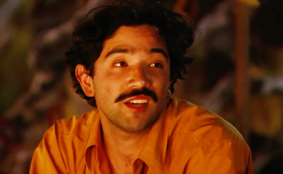An individual with dark hair and a mustache, wearing an orange shirt, smiling against a blurred, warm-toned background.