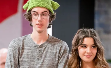 A person wearing a green knit hat with ear flaps stands next to a smiling individual with wavy hair indoors.