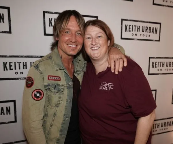 "Person in a denim jacket posing with a fan at a 'Keith Urban on Tour' event backdrop, both smiling."