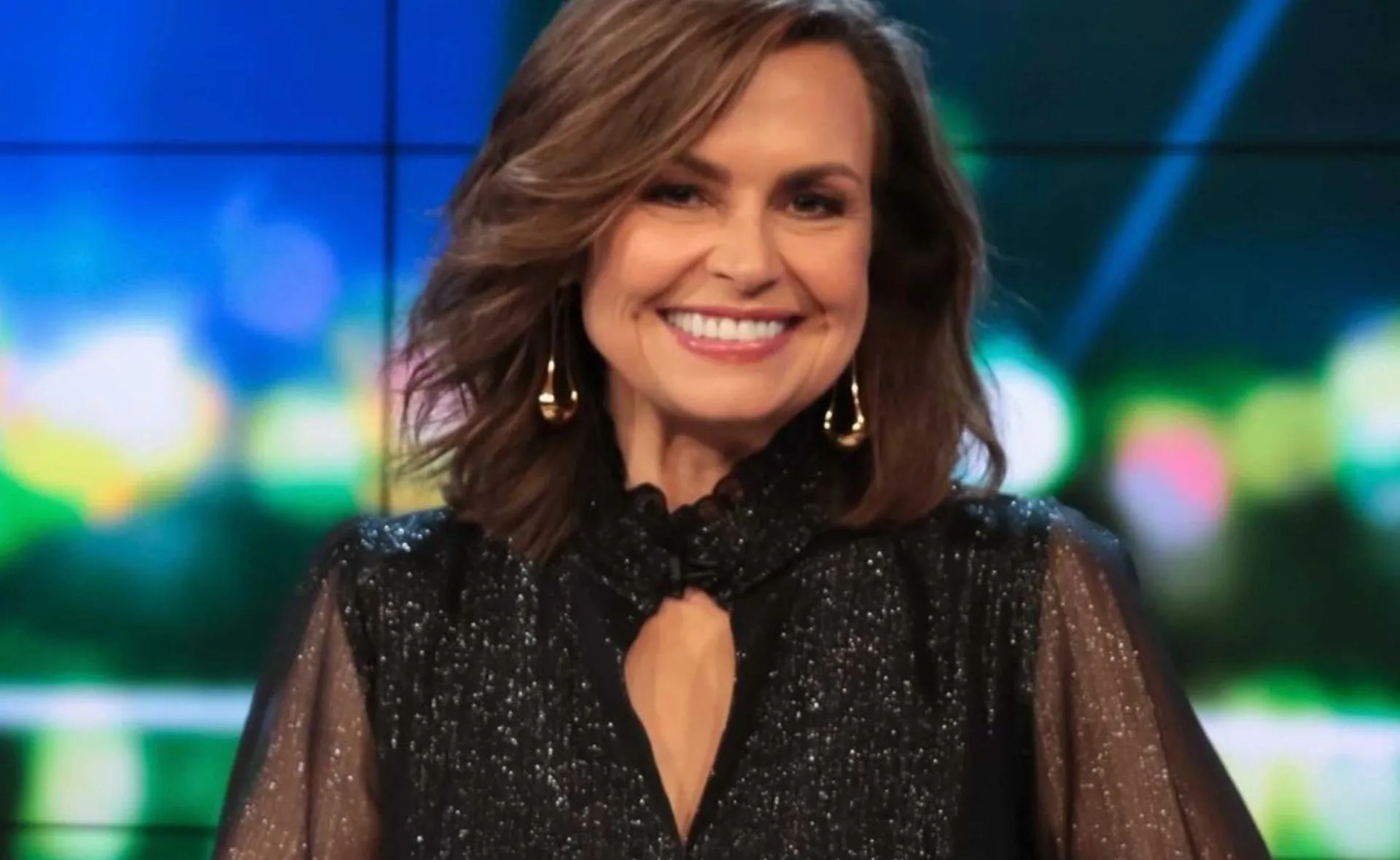 Smiling woman with wavy hair in a black glittery top and earrings, against a colorful blurred background.