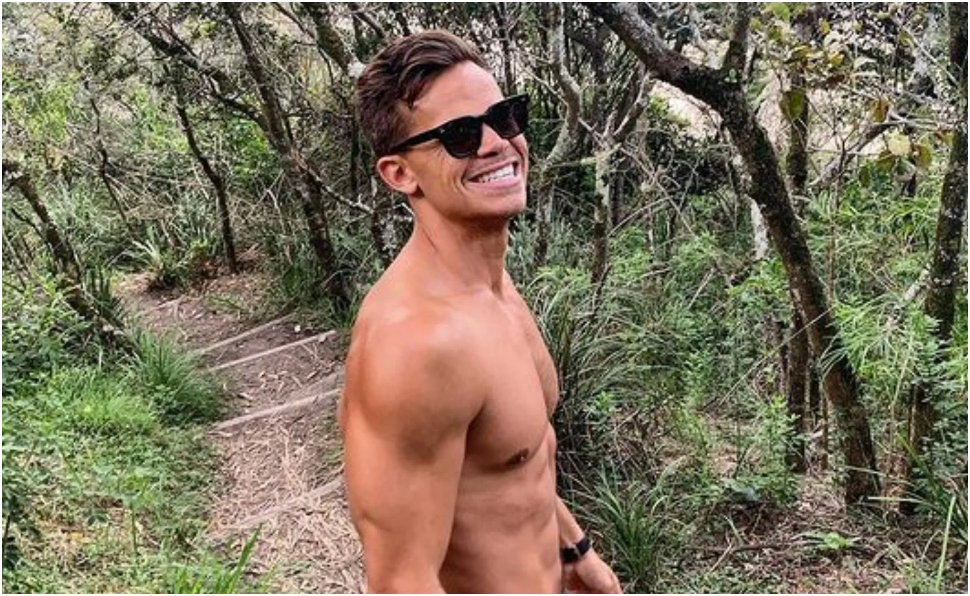Man in sunglasses, shirtless, smiling on a forest trail surrounded by greenery.