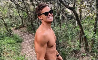 Man in sunglasses, shirtless, smiling on a forest trail surrounded by greenery.