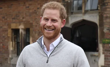 A man with red hair and a beard, smiling outdoors in front of a brick building wearing a gray sweater.