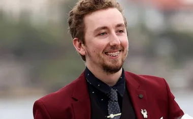 Man in a maroon suit smiling outdoors with a blurred background.