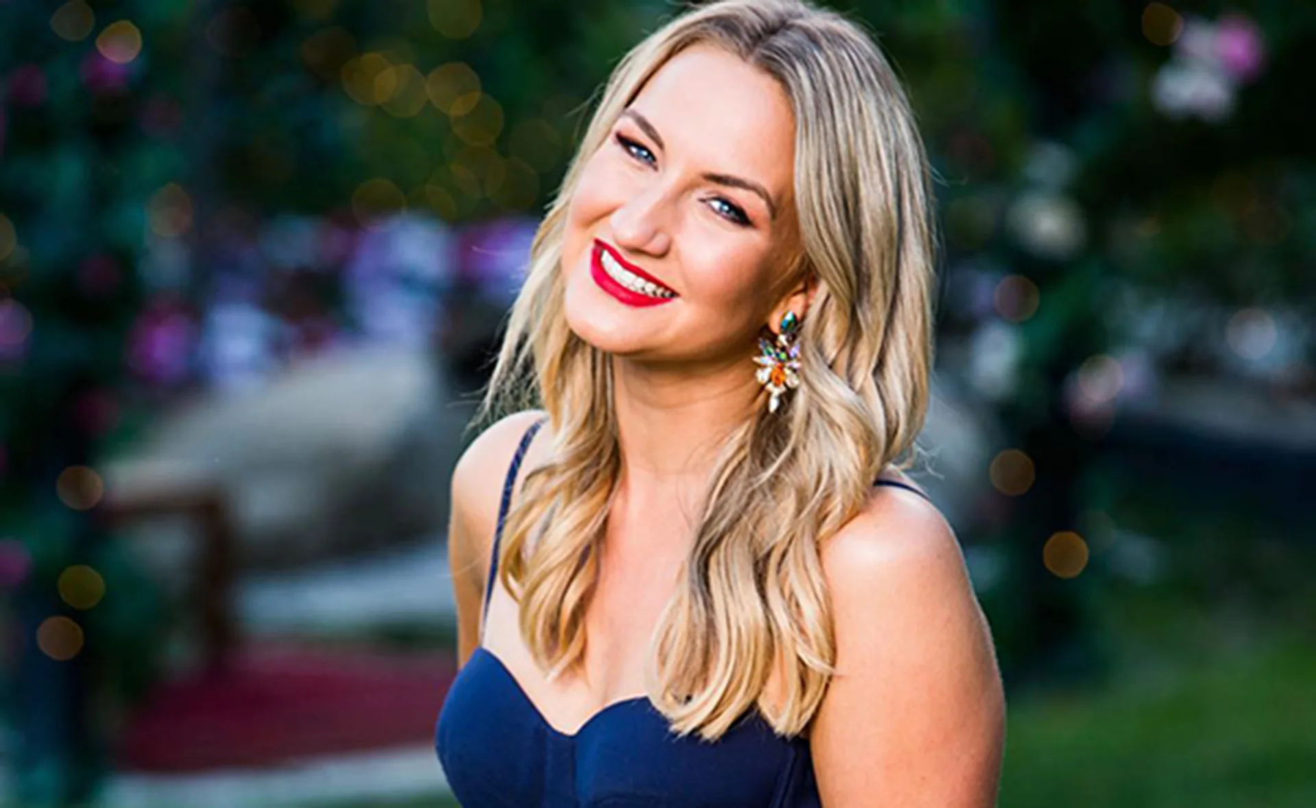 Smiling woman with wavy blonde hair, wearing a dark blue dress and colorful earrings against a blurred outdoor backdrop.