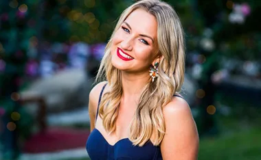 Smiling woman with wavy blonde hair, wearing a dark blue dress and colorful earrings against a blurred outdoor backdrop.