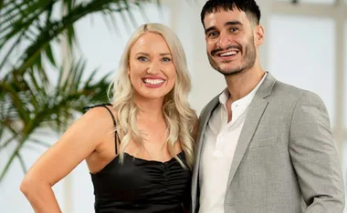 A smiling woman in a black dress stands next to a man in a gray blazer and white shirt, with a leafy background.