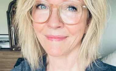 A person with short blonde hair and large glasses smiling at the camera indoors.