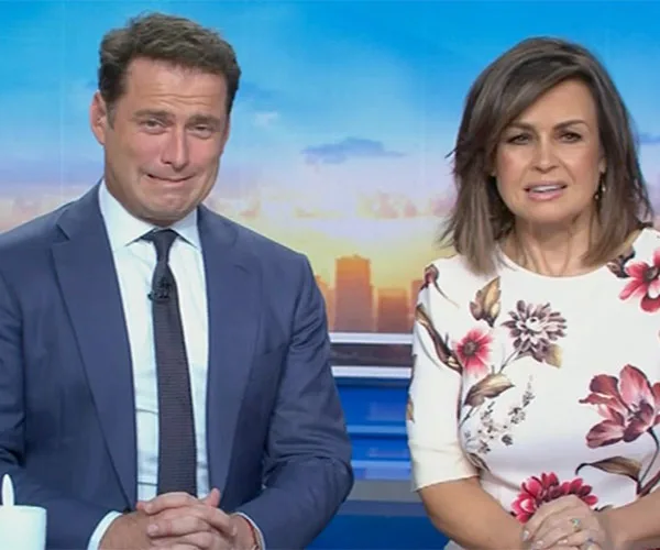 Two TV presenters seated, one in a suit and tie, the other in a floral dress, in a studio setting with a sunrise backdrop.