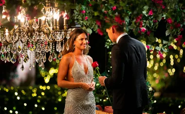 A woman in a sparkling dress receives a red rose from a man in a suit under a chandelier decorated with flowers.