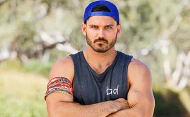 Man wearing a blue cap and sleeveless shirt, arms crossed, looking serious, standing outdoors with blurred background.