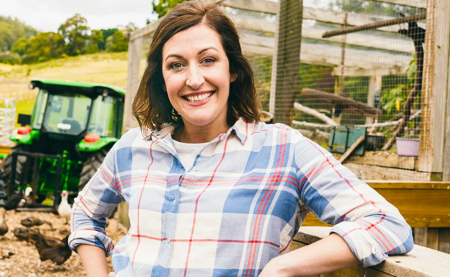 Woman in a plaid shirt smiles on a farm, with a green tractor and chickens in the background.