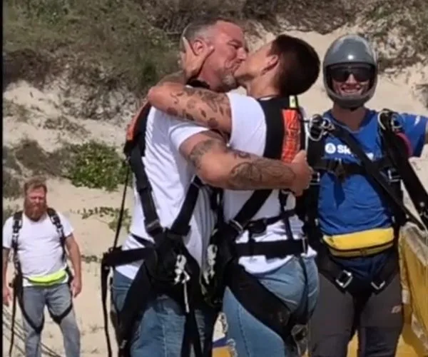 Couple kissing in skydiving gear on a sandy landscape, with parachute instructor smiling in the background.