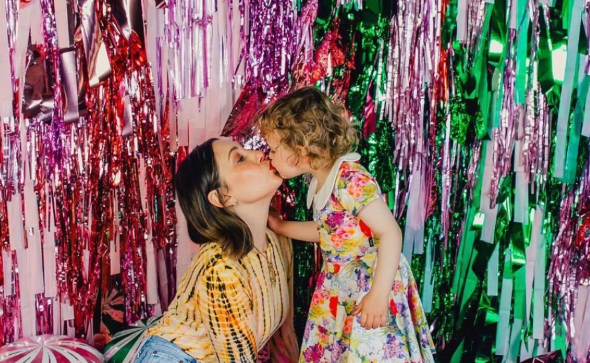 A woman and child share a kiss in front of colorful streamers, the child in a floral dress.