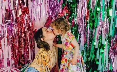 A woman and child share a kiss in front of colorful streamers, the child in a floral dress.