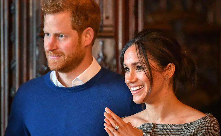 A smiling couple stands together; the woman claps while showing an engagement ring.