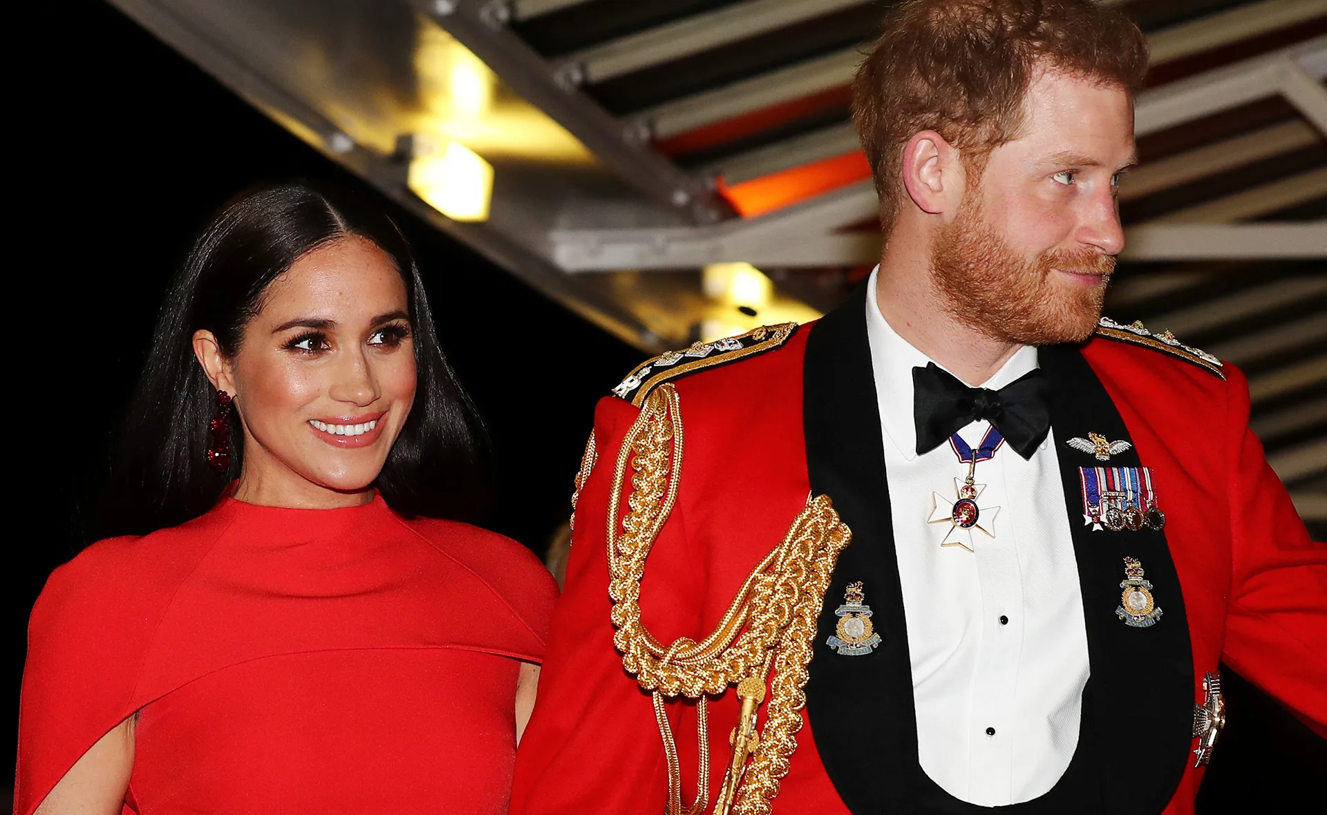 Prince Harry in military uniform with Meghan Markle in a red dress, smiling at an event.