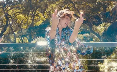 Woman in floral outfit poses with middle fingers up, surrounded by sparkling light and greenery.