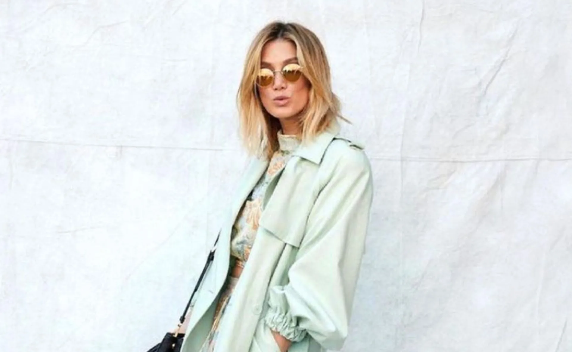 A woman in sunglasses and a light green coat poses against a plain backdrop, exuding a stylish and confident aura.