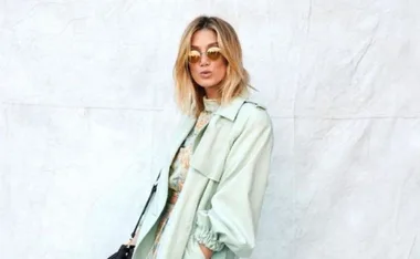 A woman in sunglasses and a light green coat poses against a plain backdrop, exuding a stylish and confident aura.