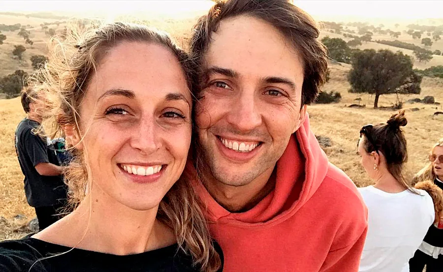 A smiling couple takes a selfie outdoors in a field, with people and trees in the background.