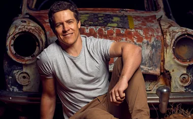 Man in a gray t-shirt and tan pants smiling, sitting in front of a rusty old car.