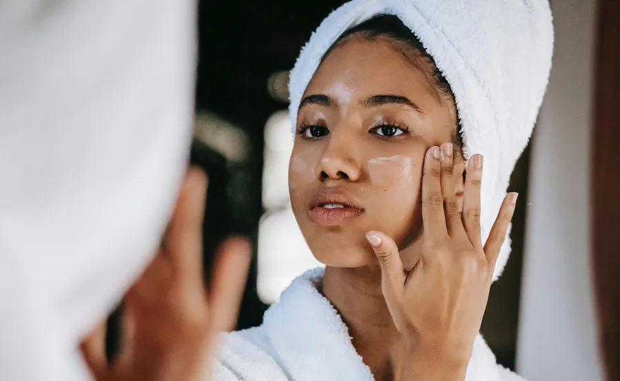 Woman in a towel applying cream to her face while looking in the mirror.