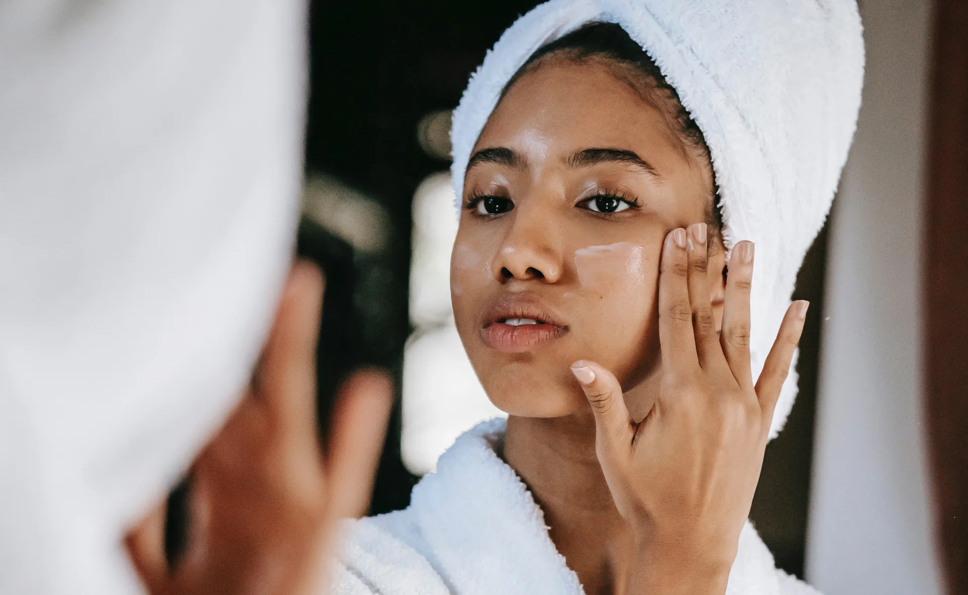 Woman in a towel applying cream to her face while looking in the mirror.