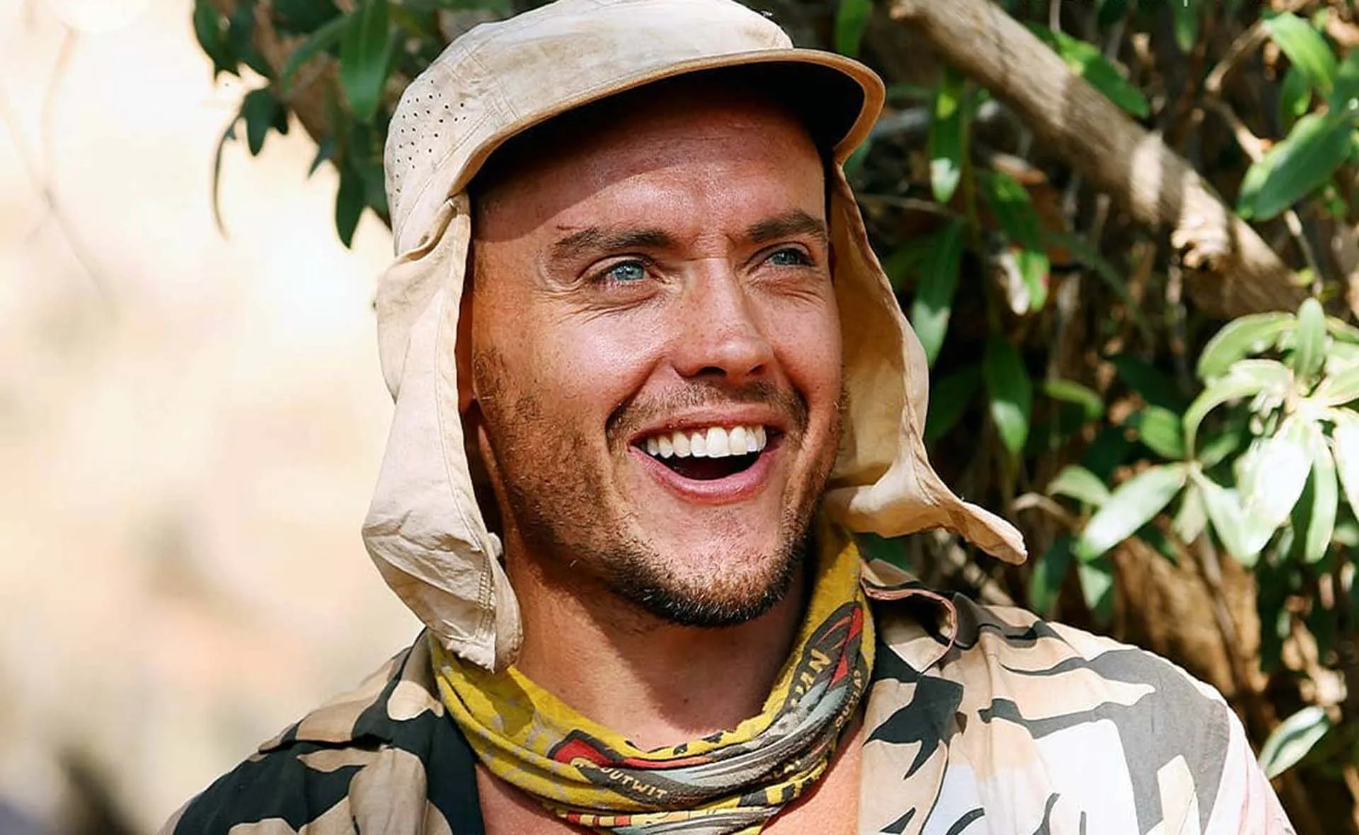 Man smiling in outdoor setting, wearing a beige hat and patterned scarf around neck.
