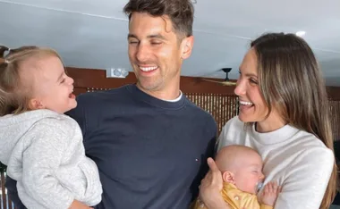 A couple smiling and holding their two young children, one toddler and one baby, in a living room setting.