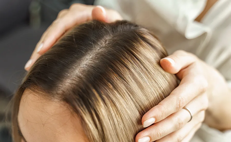 Person applying gentle pressure on a woman's scalp, showcasing brown roots and blonde hair, possibly during home haircare.