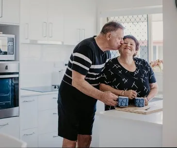Couple in a kitchen, man kisses woman on cheek, both holding mugs on counter.