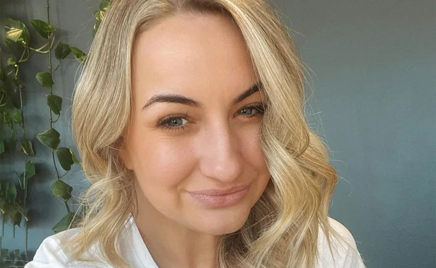 Blonde woman smiling softly, with wavy hair and green plants in the background.