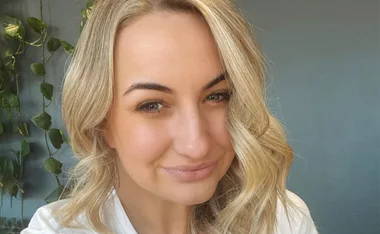 Blonde woman smiling softly, with wavy hair and green plants in the background.