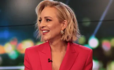 Woman in a red jacket smiling during a TV appearance, with a blurred colorful background.