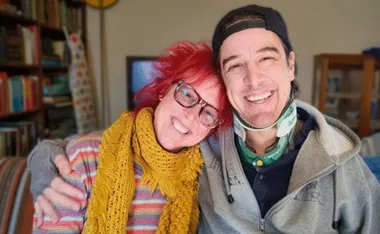 A smiling couple, one with red hair and glasses, the other wearing a neck brace and cap, embraces in a cozy room.