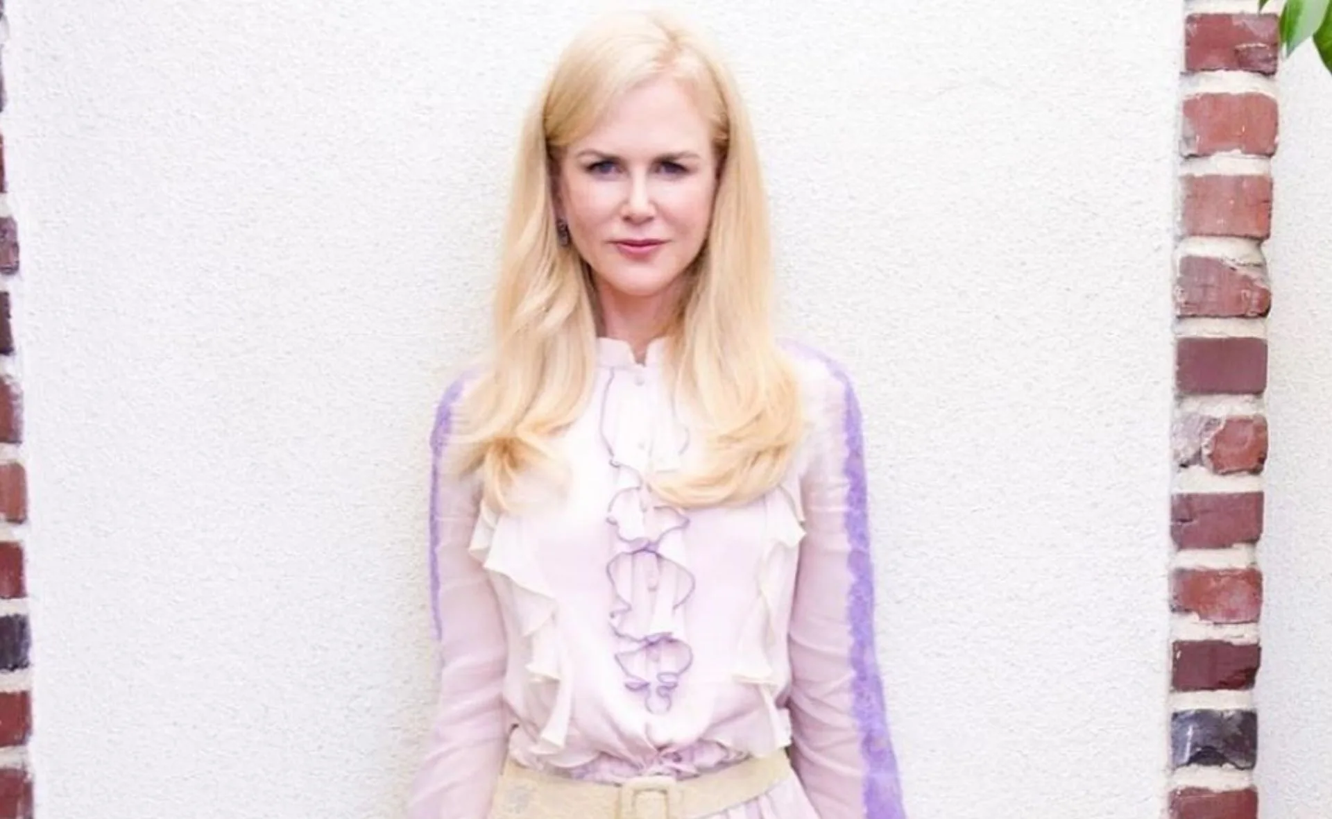 Blonde woman with straight hair in a pink and purple frilled blouse, standing against a white and brick background.